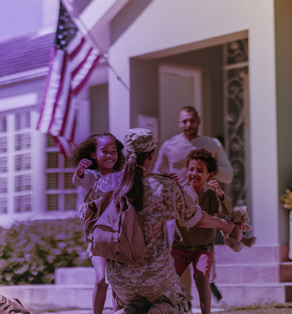 A U.S. military service member kneeling and embracing two joyful children running toward them outside a home.