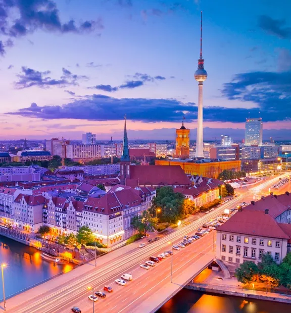 Panoramic aerial view of Berlin, Germany, at sunset, featuring the Spree River, historic buildings, illuminated streets, and the city skyline.