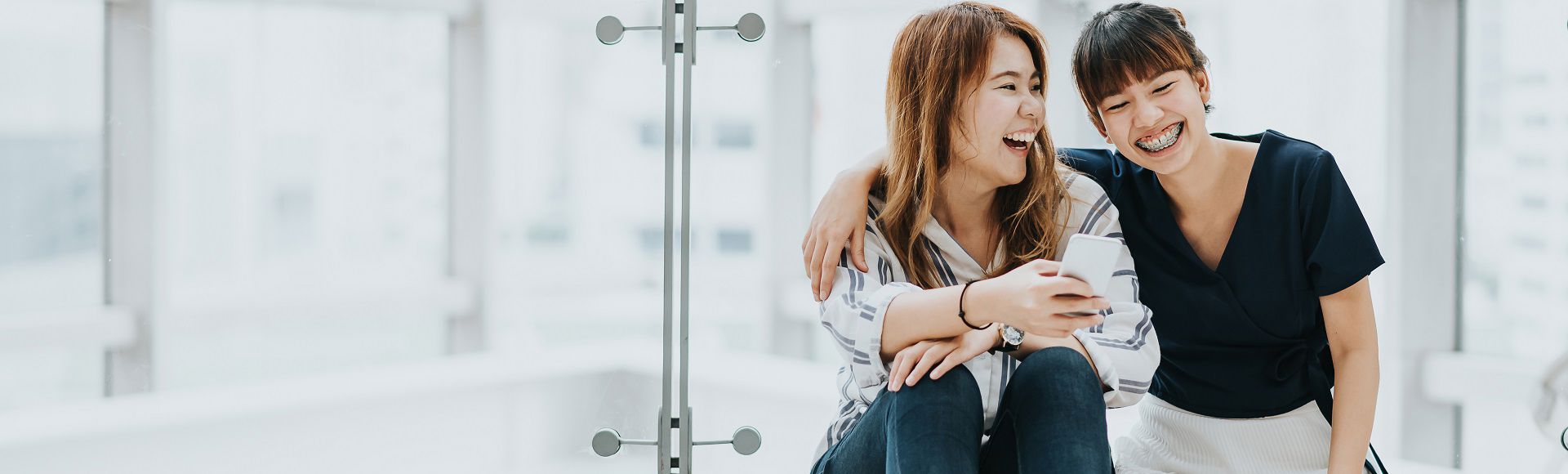 Two friends sitting together indoors, laughing while looking at a smartphone.