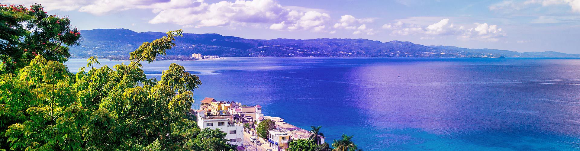 Panoramic view of Montego Bay, Jamaica, overlooking the turquoise Caribbean Sea, coastal buildings, lush tropical vegetation, and mountains in the distance.