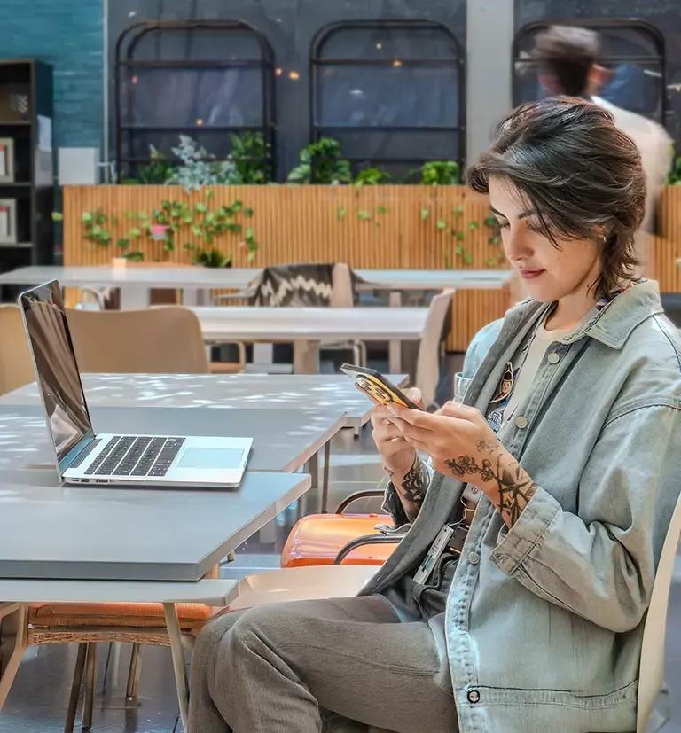 Woman using a smartphone beside a laptop in a modern workspace.