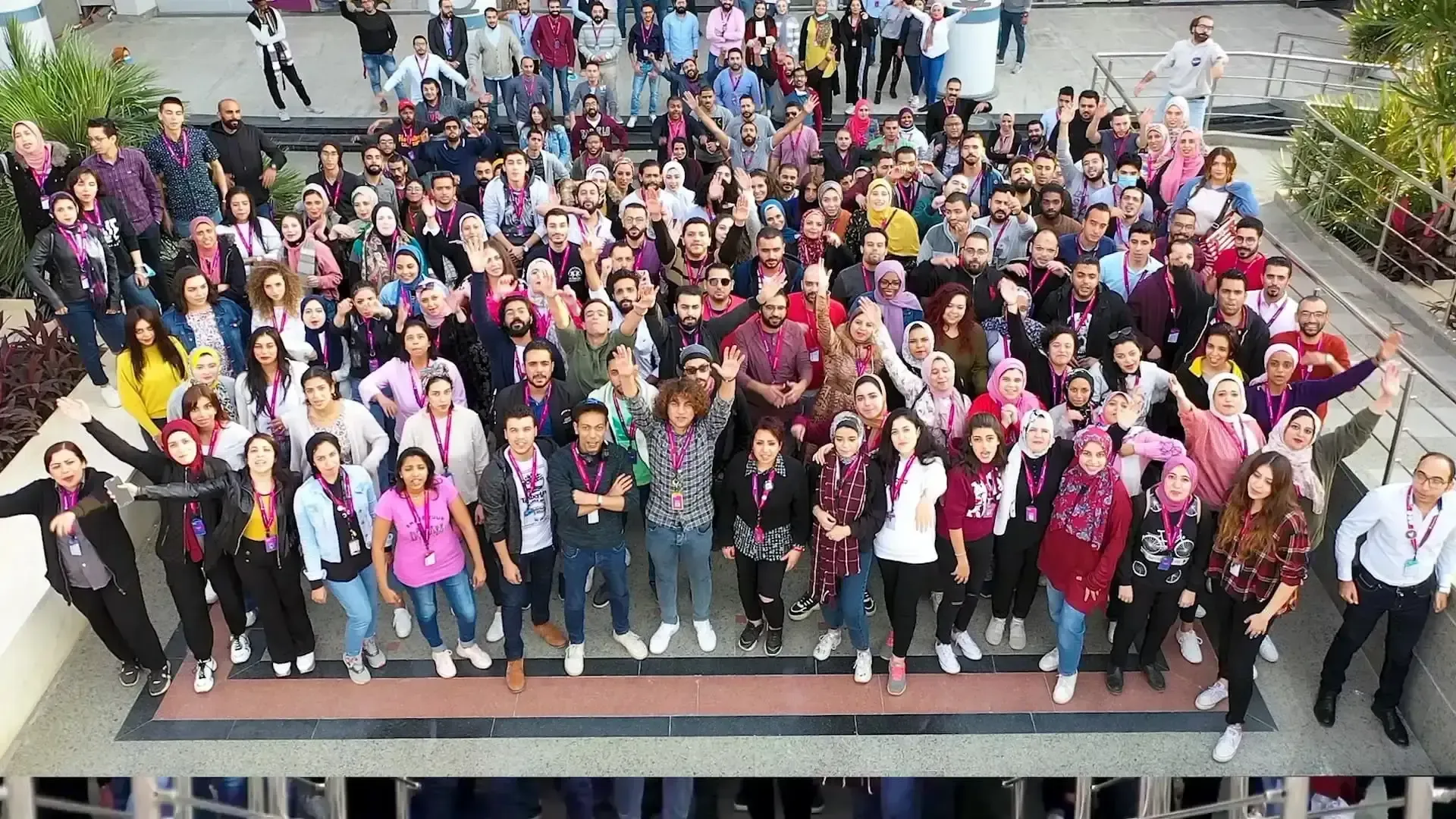 Large group of people gathered outdoors, many wearing colorful clothing and smiling while waving at the camera.