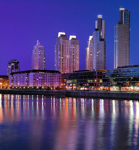 Nighttime view of Buenos Aires, Argentina, featuring modern high-rise buildings, illuminated waterfront restaurants, and colorful reflections on the water.