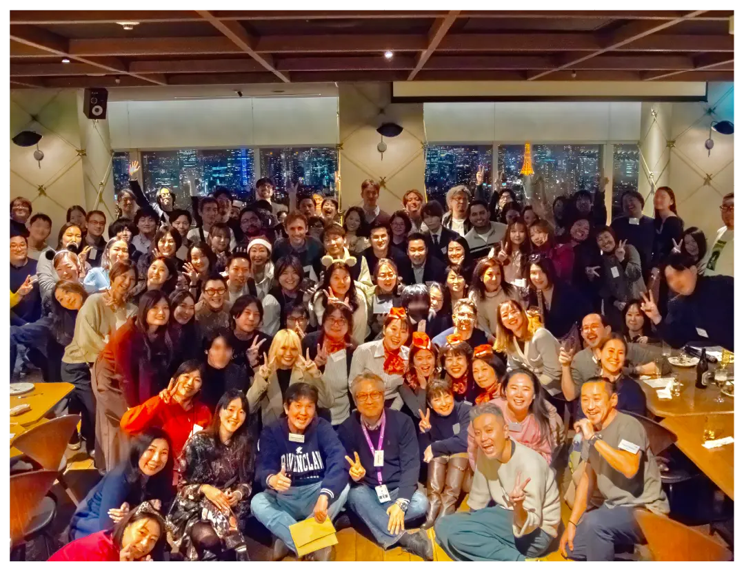 A large diverse group poses joyfully indoors with a Tokyo night view.