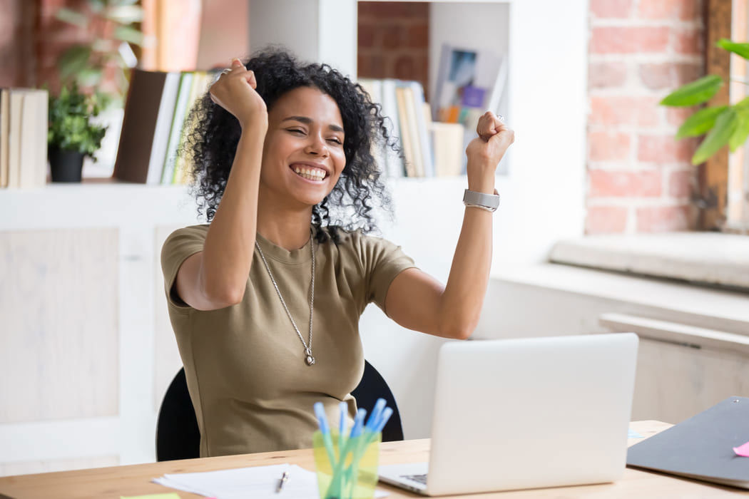Afro-descendant woman celebrating with raised arms while working on her computer.