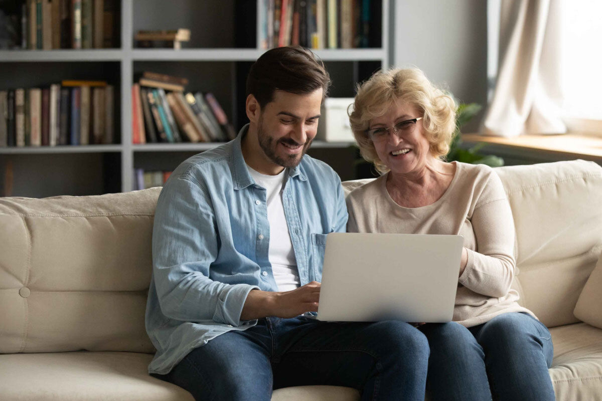 A young man and an elderly lady sit together on a sofa looking at a laptop.