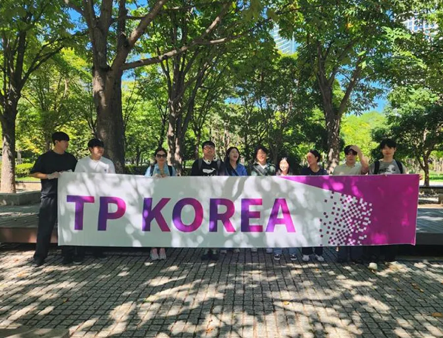 TP Korea team members pose together holding a branded banner in a sunny park.