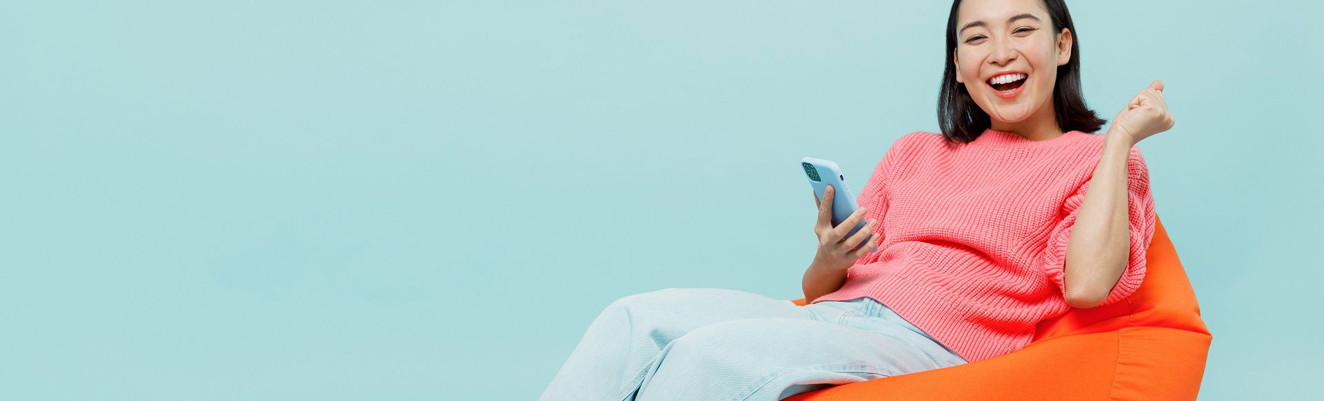 Woman smiling while holding a smartphone and celebrating on an orange chair.