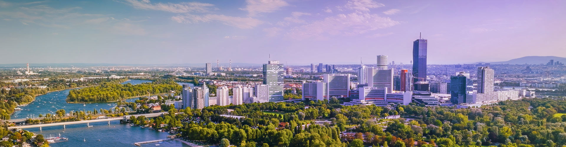 Panoramic aerial view of Vienna, Austria, featuring the modern city skyline, the Danube River, bridges, and lush green parks.