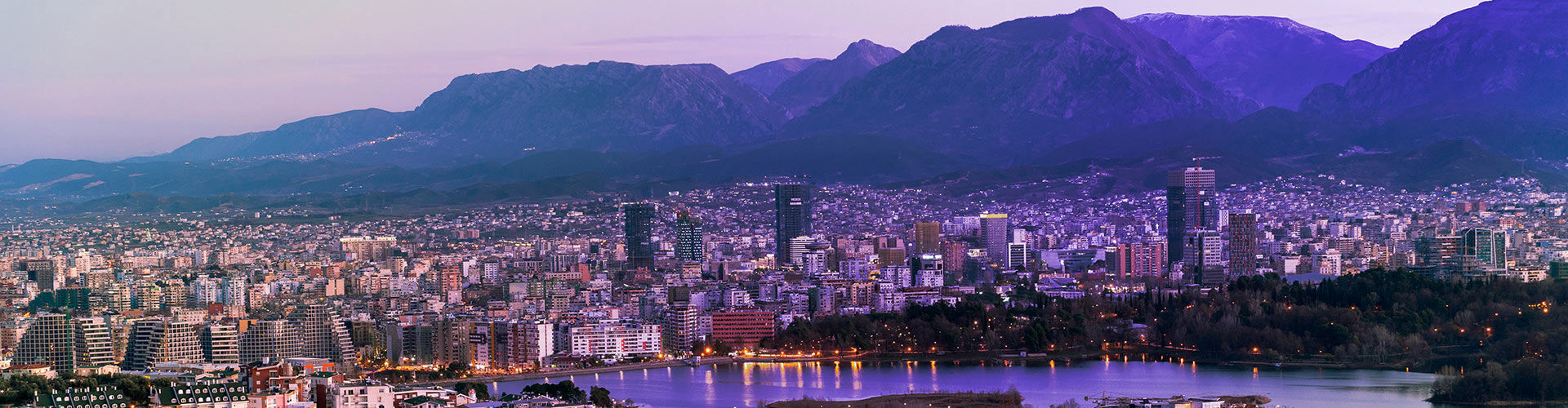 Panoramic view of Tirana, Albania, at dusk, featuring a dense city skyline, modern buildings, a waterfront park, and mountains in the background.