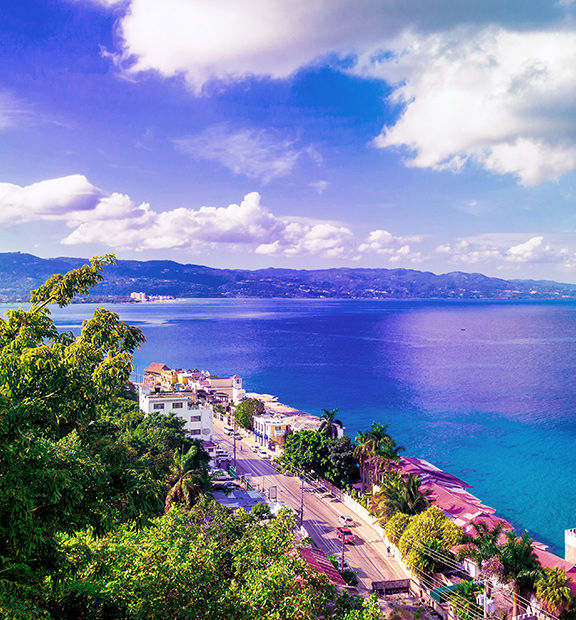 Panoramic view of Montego Bay, Jamaica, overlooking the turquoise Caribbean Sea, coastal buildings, lush tropical vegetation, and mountains in the distance.