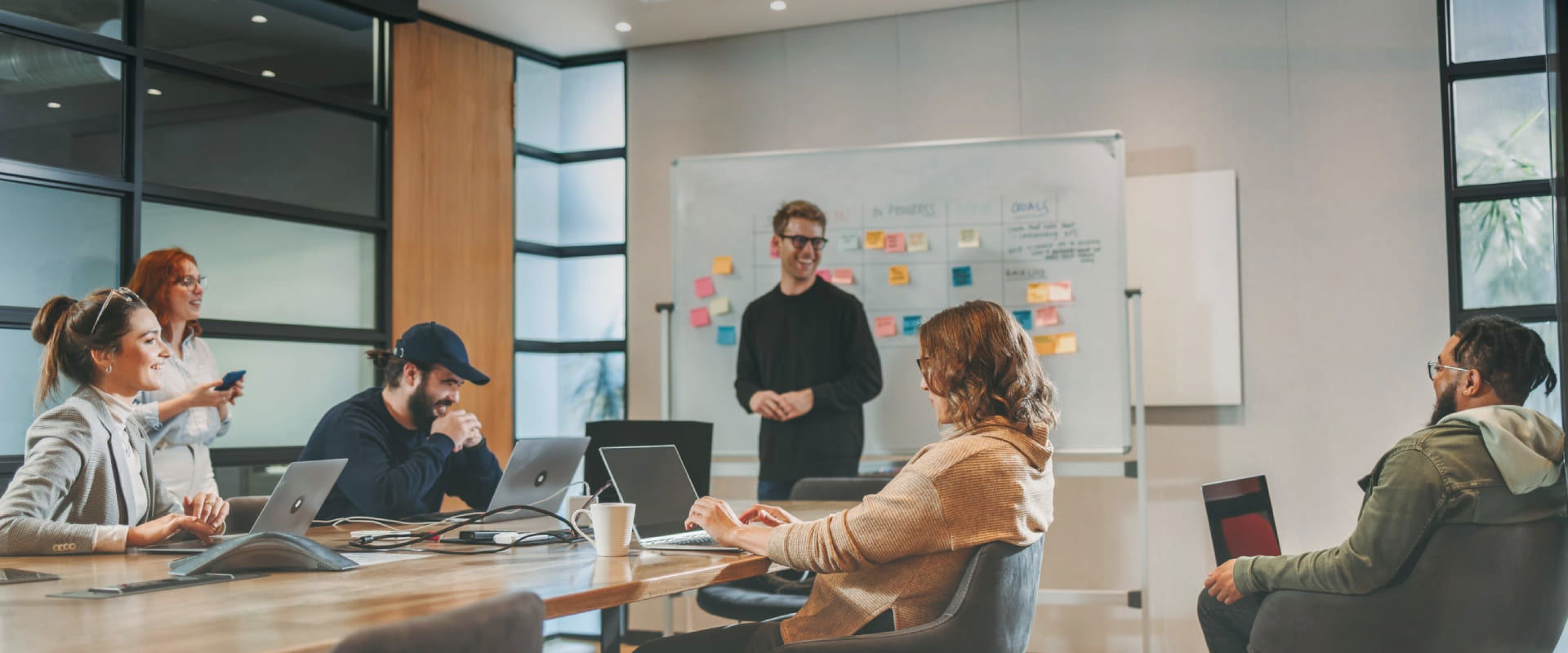 Group of people in a modern meeting room with laptops and sticky notes on the wall.
