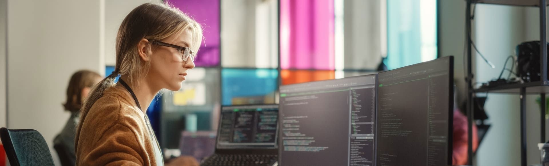 Blonde woman wearing glasses focused on coding across dual monitors.