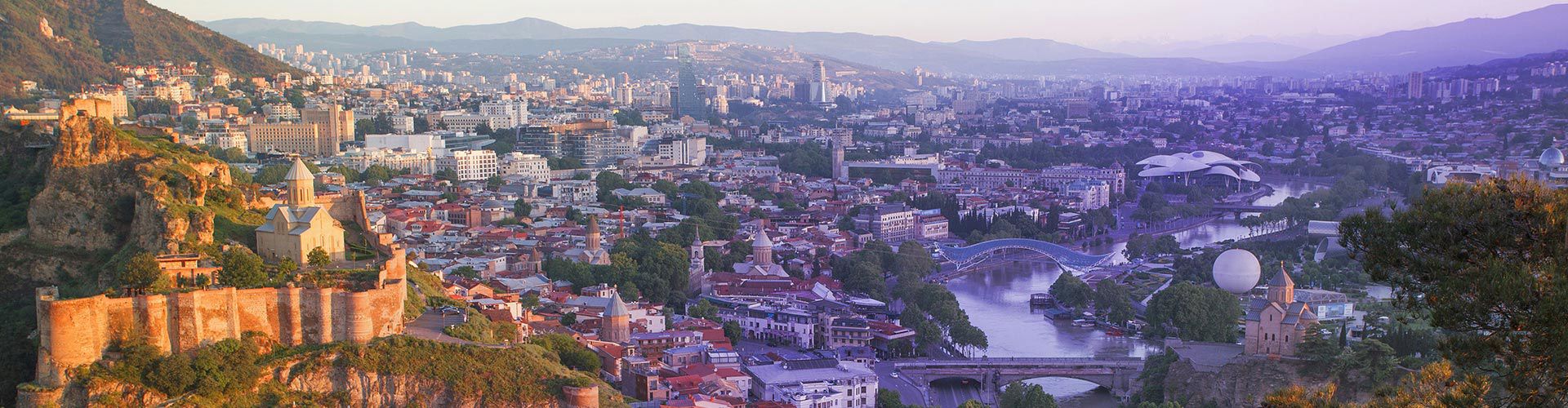 Panoramic view of Tbilisi, Georgia, featuring the historic old town, Narikala Fortress, the Kura River, modern architecture, and mountains in the background.