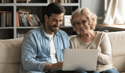 A young man and an elderly lady sit together on a sofa looking at a laptop.