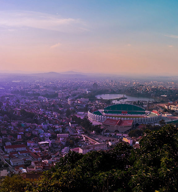 Panoramic view of Antananarivo, Madagascar, at sunset, featuring the city’s dense urban landscape, hills, a large stadium, and mountains in the distance.