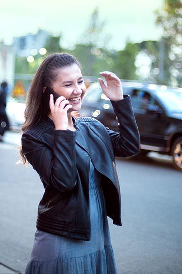 A person is happily talking on a phone, smiling, and gesturing with a hand. they are standing on a street with blurred cars and trees in the background.