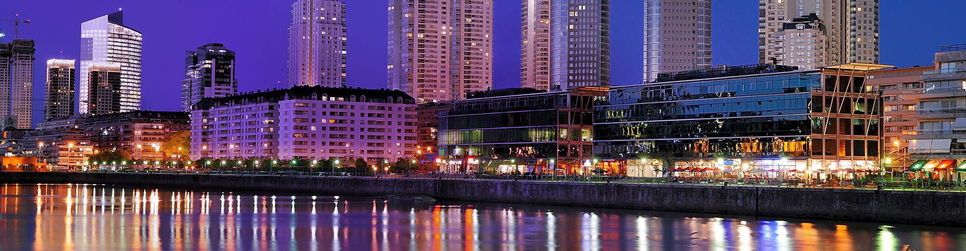 Nighttime view of Buenos Aires, Argentina, featuring modern high-rise buildings, illuminated waterfront restaurants, and colorful reflections on the water.