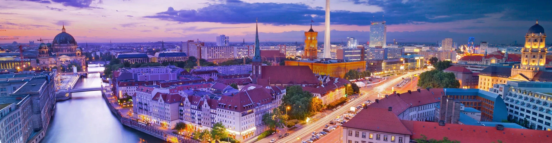 Panoramic aerial view of Berlin, Germany, at sunset, featuring the Spree River, historic buildings, illuminated streets, and the city skyline.