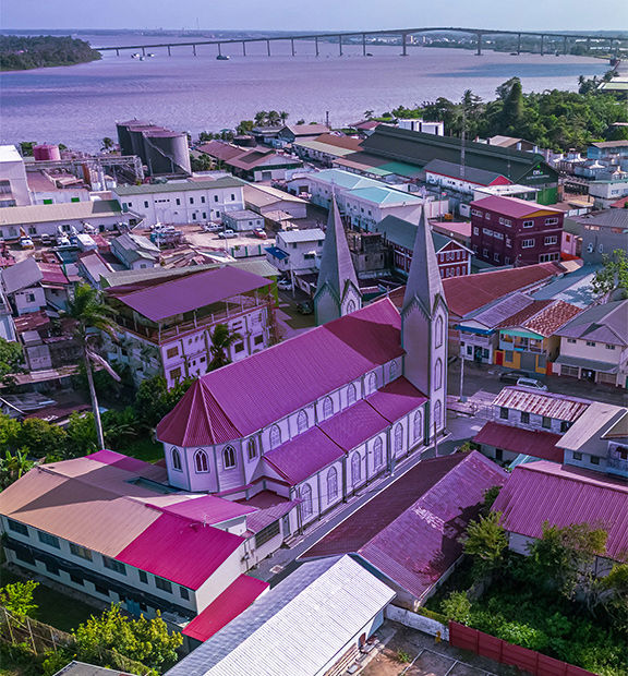 Aerial view of Paramaribo, Suriname, featuring a church with a tall steeple surrounded by colorful rooftops, residential buildings, tree-lined streets, and a waterfront area.