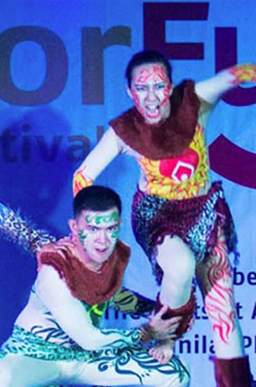 Two male dancers in vibrant tribal costumes perform on a stage with blue lighting.