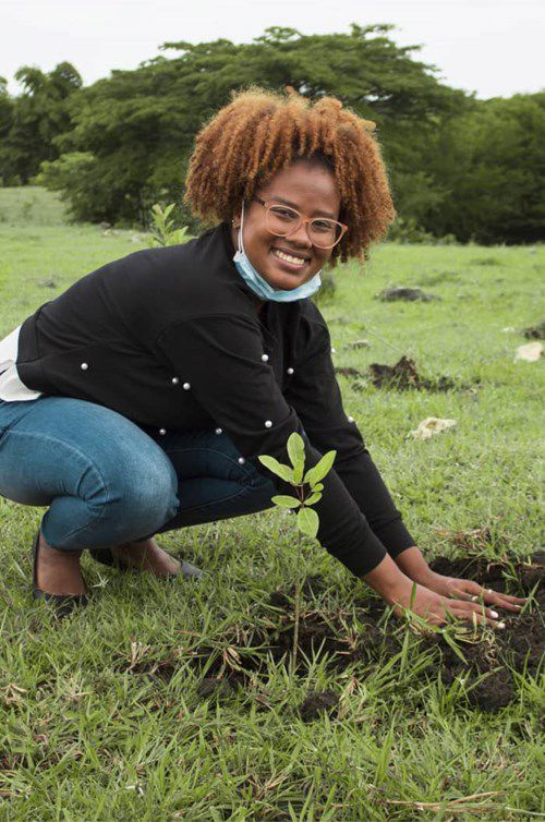 a person crouches while planting a small sapling in a grassy field, smiling and wearing a black jacket and a face mask under their chin; trees are visible in the background.