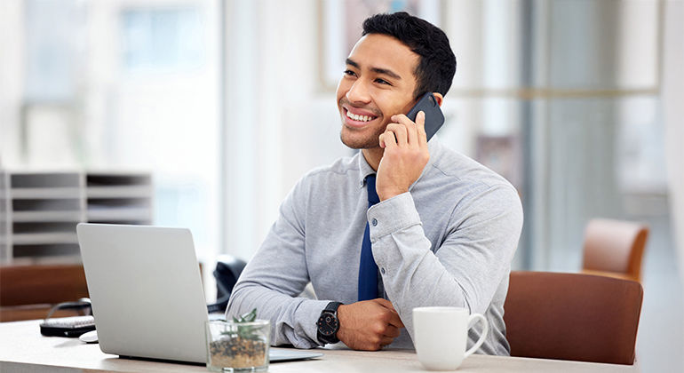 Business professional smiling during a phone call at a modern office desk with laptop, coffee mug, and potted plant, dressed in gray shirt and blue tie.