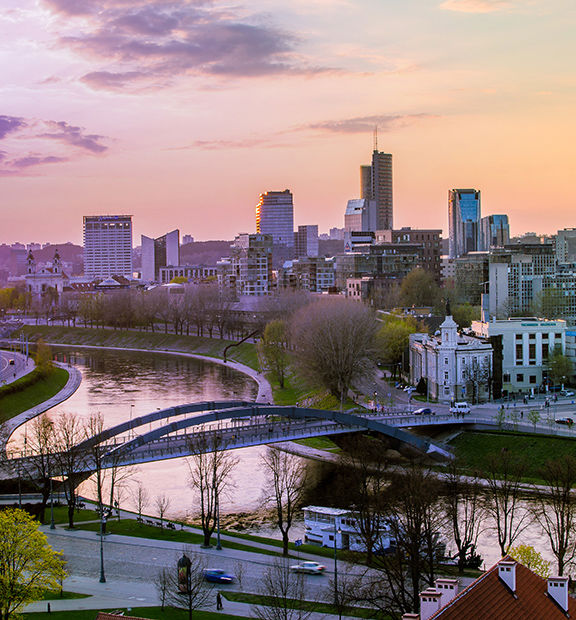 Panoramic view of Vilnius, Lithuania, at sunset, featuring the Neris River, historic buildings, tree-lined streets, and the modern city skyline.