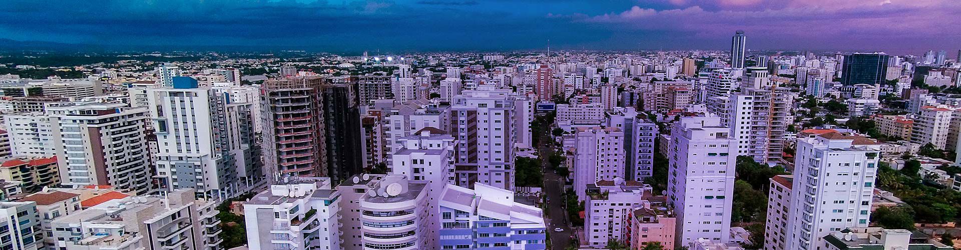Aerial panoramic view of a dense urban skyline in the Dominican Republic at dusk, showing rows of high-rise apartment and office buildings under a dramatic sky transitioning from deep blue to pink and purple clouds.